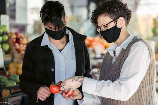 Two individuals wearing masks choose fresh tomatoes at a local market, highlighting safety and healthy choices.